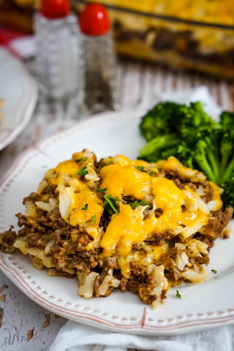 A serving of Hamburger Hashbrown Casserole is served with cooked broccoli on a white plate.