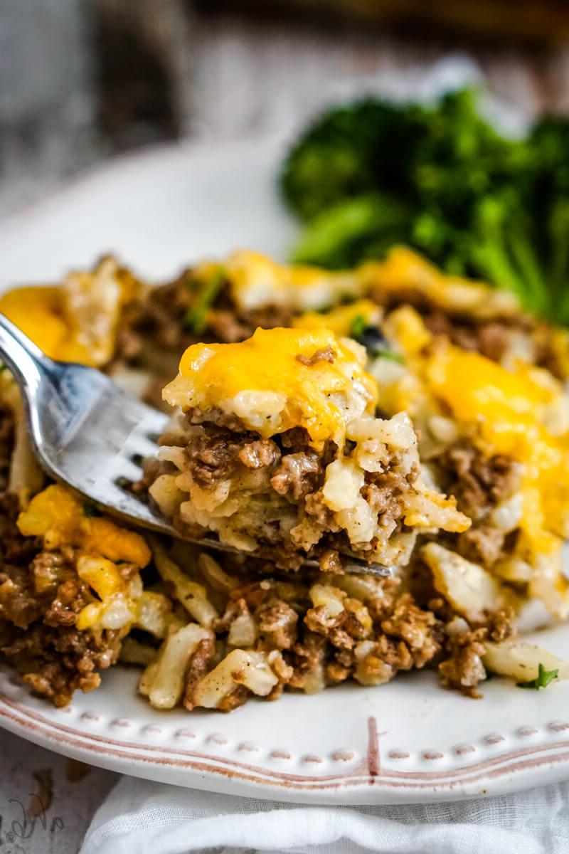 A forkful of casserole is held above a serving of casserole with steamed broccoli on a white plate.