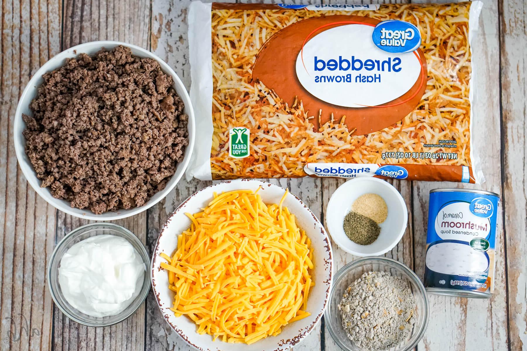 A bag of shredded hashbrowns, a bowl of browned ground beef, a bowl of shredded cheddar cheese, pepper, garlic powder, a can of cream of mushroom soup, dry onion soup mix, and sour cream.