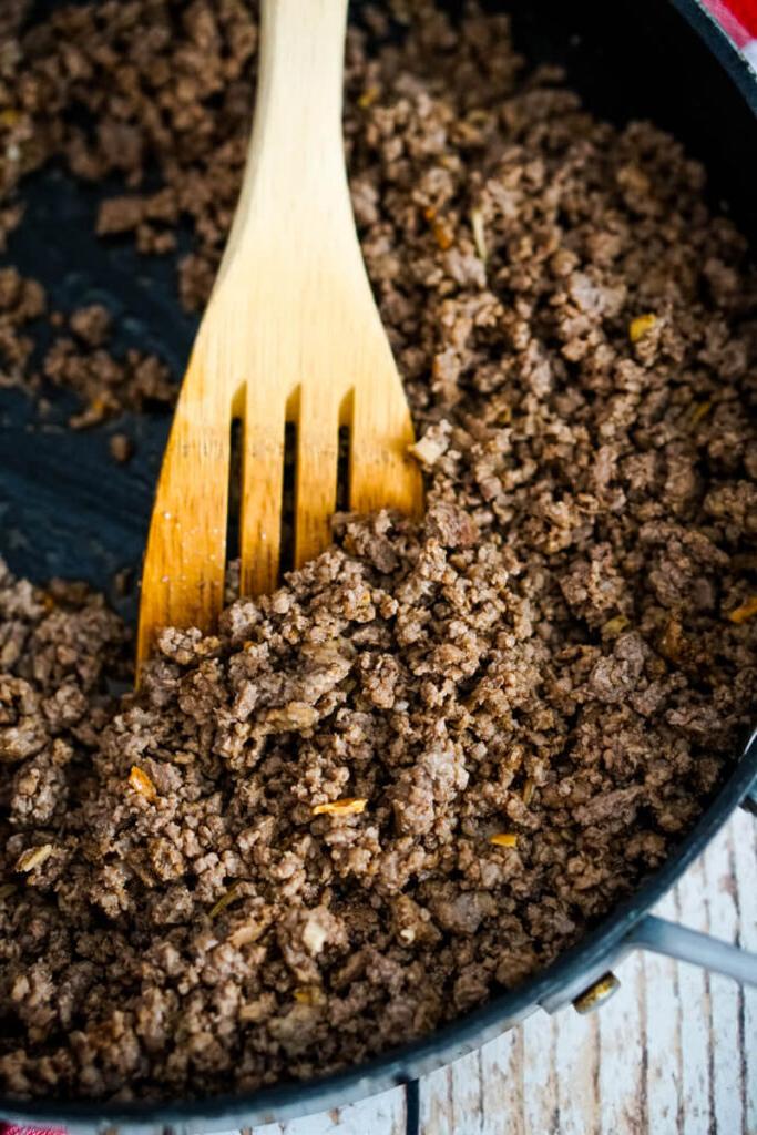 Browned ground beef is stirred by a wooden spoon in a skillet.