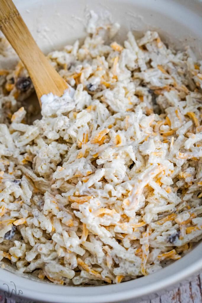 Hash browns, cheese, cream of mushroom soup, sour cream, pepper, and garlic powder are combined with a wooden spoon in a white mixing bowl.