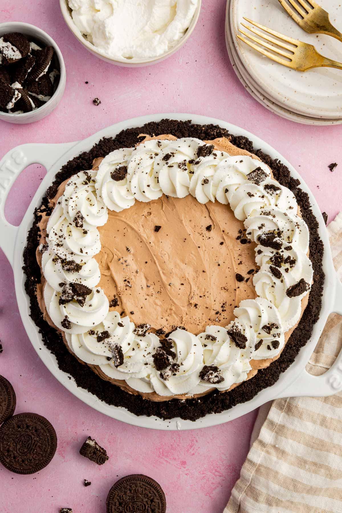 An overhead view of a gluten-free Oreo pudding pie topped with whipped cream swirls and crushed Oreos on a pink table.