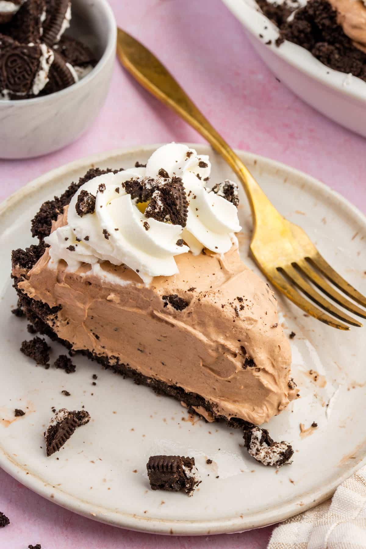 A slice of gluten-free Oreo pudding pie topped with whipped cream swirls on a dessert plate with a gold fork.