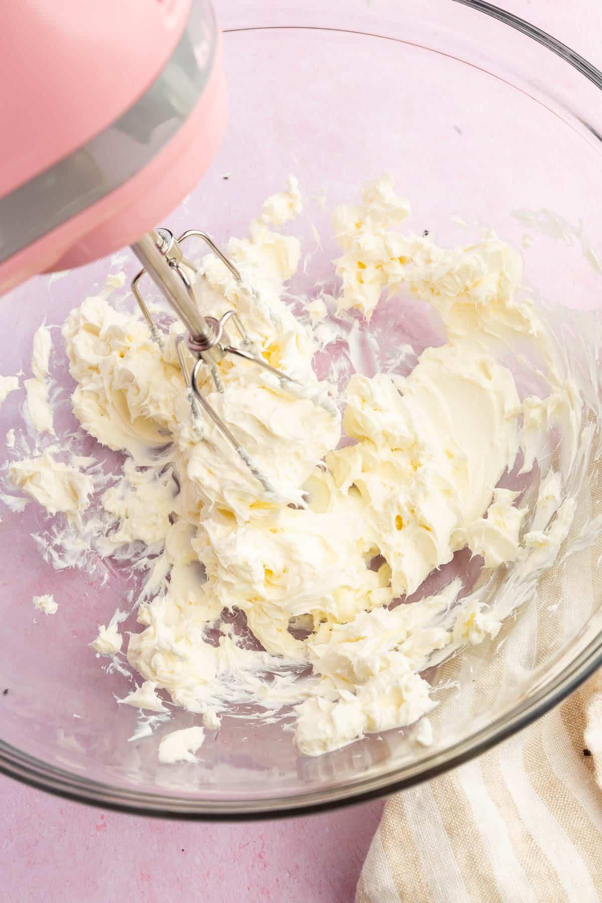 An electric hand mixer blending cream cheese in a glass mixing bowl.