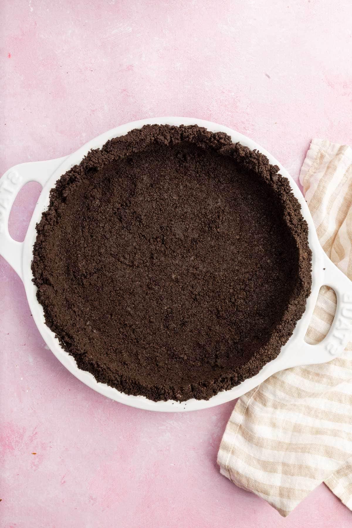 An overhead view of a gluten-free Oreo pie crust in a white pie plate on a pink table.