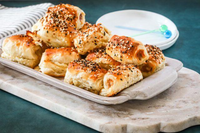 A serving tray with baked puff pastry pigs in a blanket with small appetizer plates and picks for serving.