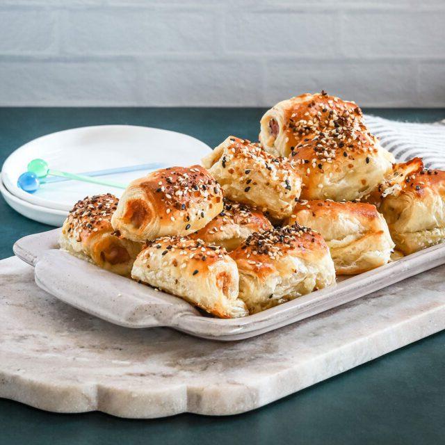 A serving tray with baked puff pastry pigs in a blanket with small appetizer plates and picks for serving.